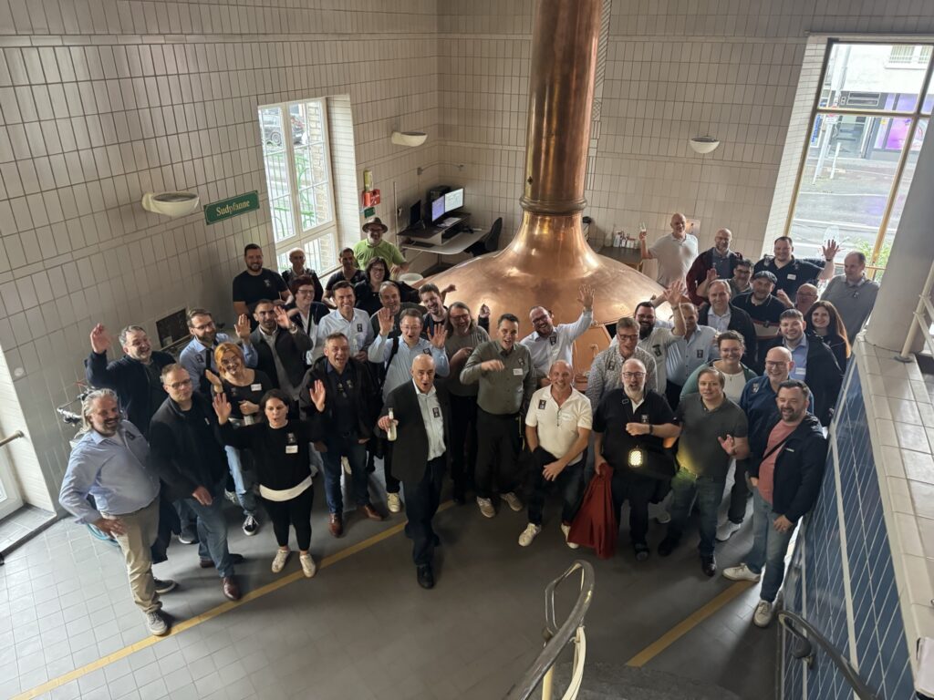 Gruppenfoto der Teilnehmenden in der BRAUWELT Köln vor dem kupfernen Sudkessel, viele winken in die Kamera.