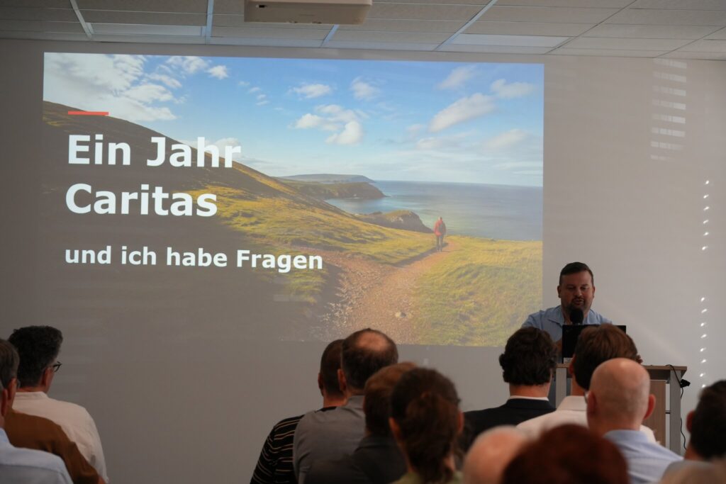 Ben Fehler am Rednerpult bei seiner Keynote, auf der Leinwand der Titel „Ein Jahr Caritas und ich habe Fragen“.